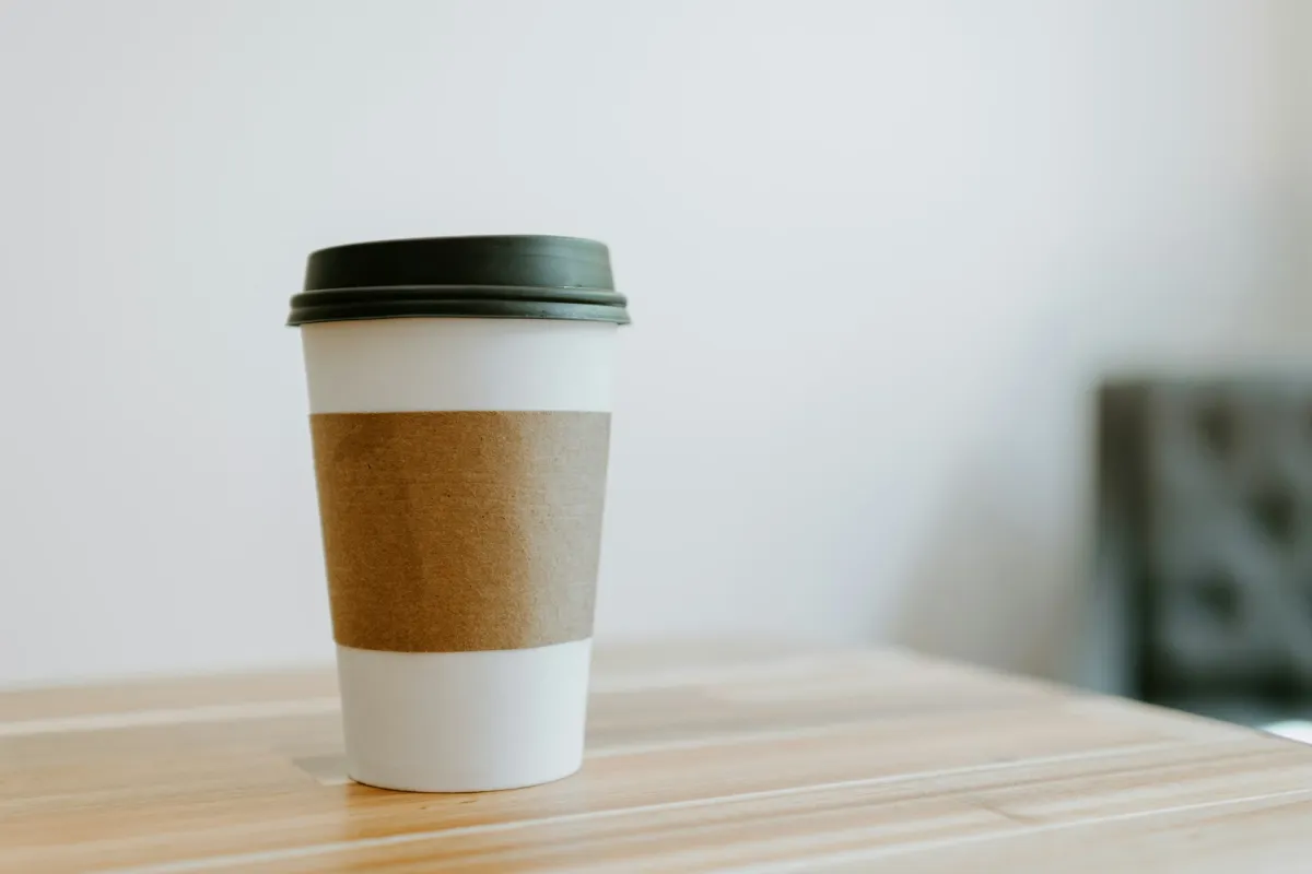 A white takeaway coffee cup with a black lid and a brown kraft sleeve on a wooden table.