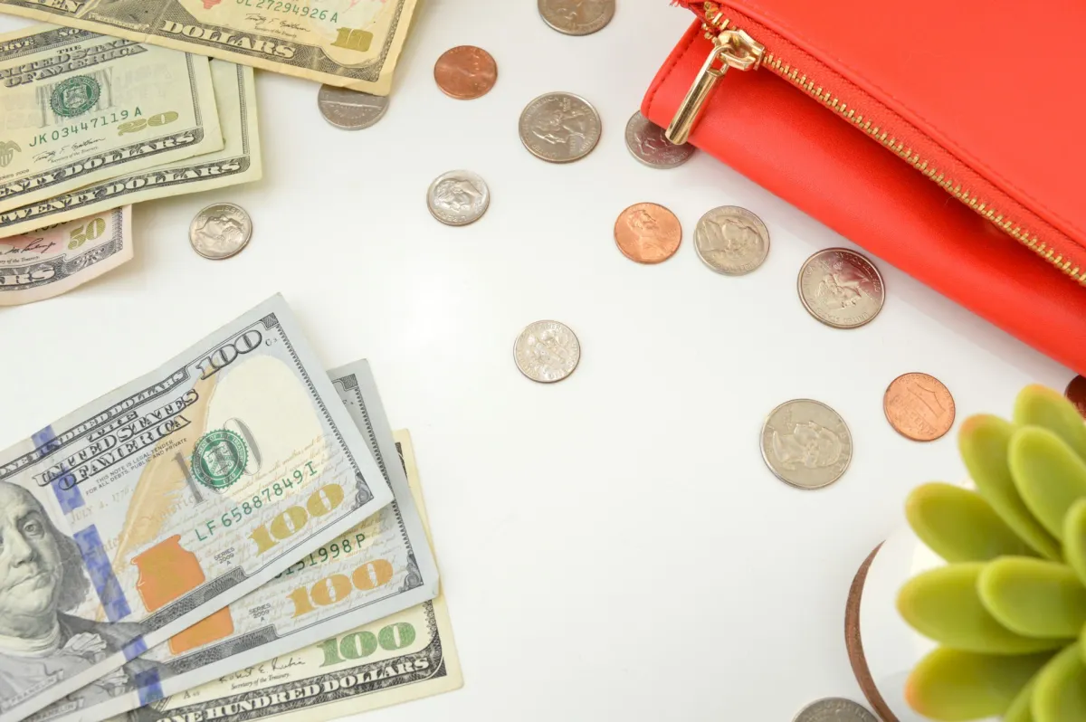 US dollar bills and scattered coins next to a red wallet and a small succulent on a white surface.