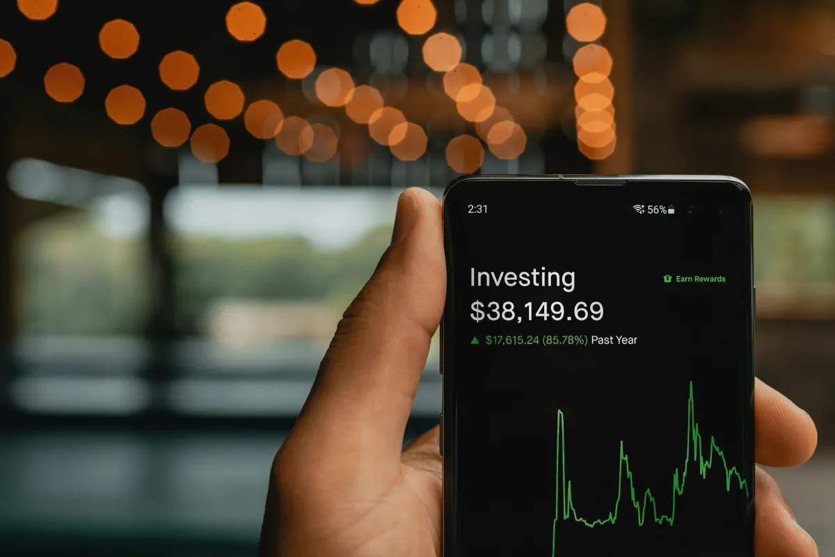 Hand holding a smartphone showing an investing app, with a portfolio balance of $38,149.69 up 85.78% over the past year on a green chart